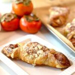 Cinnamon Persimmon Danish Twist Dessert on a white plate with 3 persimmons on top of each other in background with a silver tray with baked danishes and white canisters in background