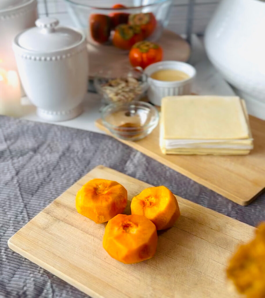 Cinnamon Persimmon Danish Twist Dessert on a white plate with 3 persimmons pealed on a wooden chopping board puff pastry in background with a grey table cloth a white and glass bowl of ingredients white canisters in background