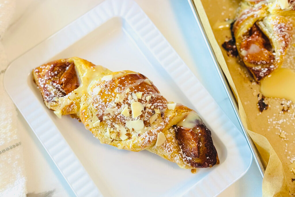 Cinnamon Persimmon Danish Twist Dessert on a white plate with 3 persimmons on top of each other in background with a silver tray with baked danishes and white canisters in background