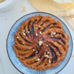 Muhamarra Roasted Red Pepper Dip in a round mezza platter with blue and white strips on top of a white countertop with a white marble mortar filled with crushed walnuts and pita bread on top of a wooden chopping board.