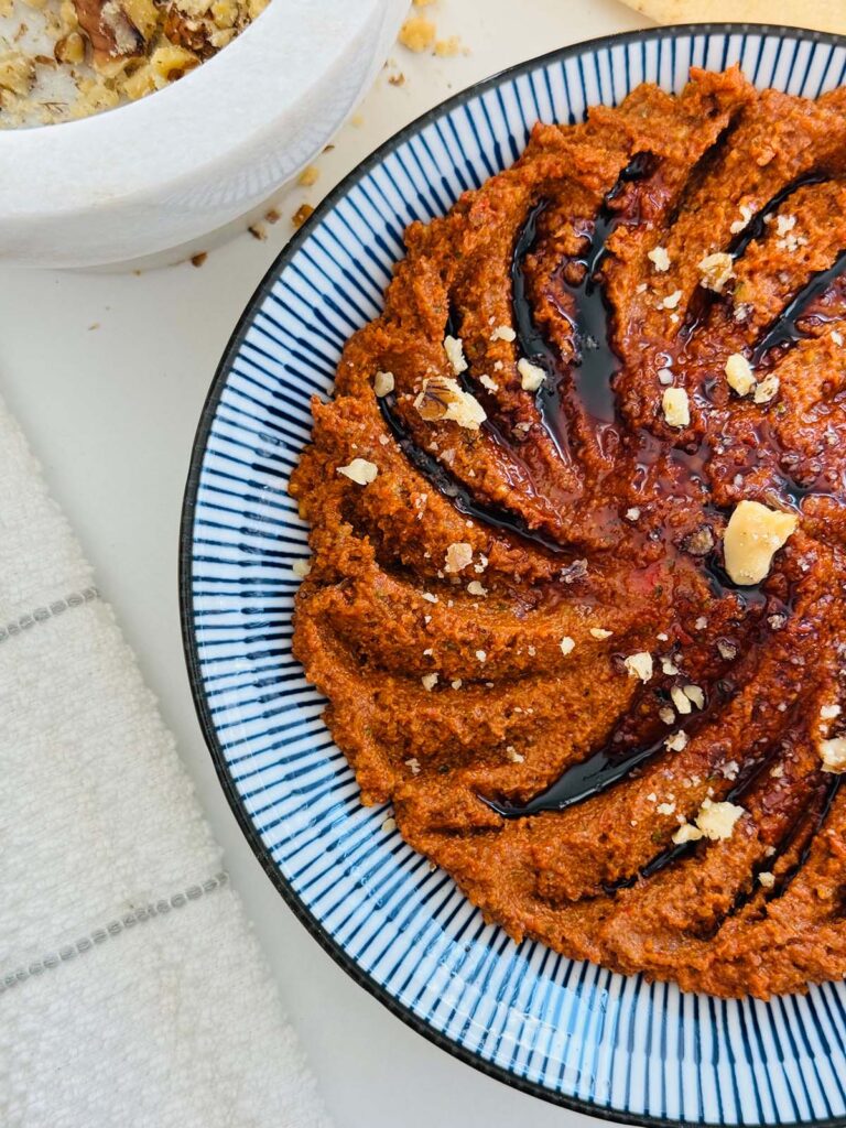 Muhamarra Roasted Red Pepper Dip in a round mezza platter with blue and white strips on top of a white countertop with a white marble mortar filled with crushed walnuts and pita bread on top of a wooden chopping board.