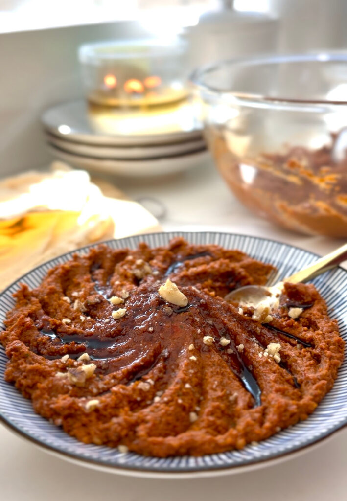 Muhamarra Roasted Red Pepper Dip in a round mezza platter with blue and white strips on top of a white countertop with a white marble mortar filled with crushed walnuts and pita bread on top of a wooden chopping board.