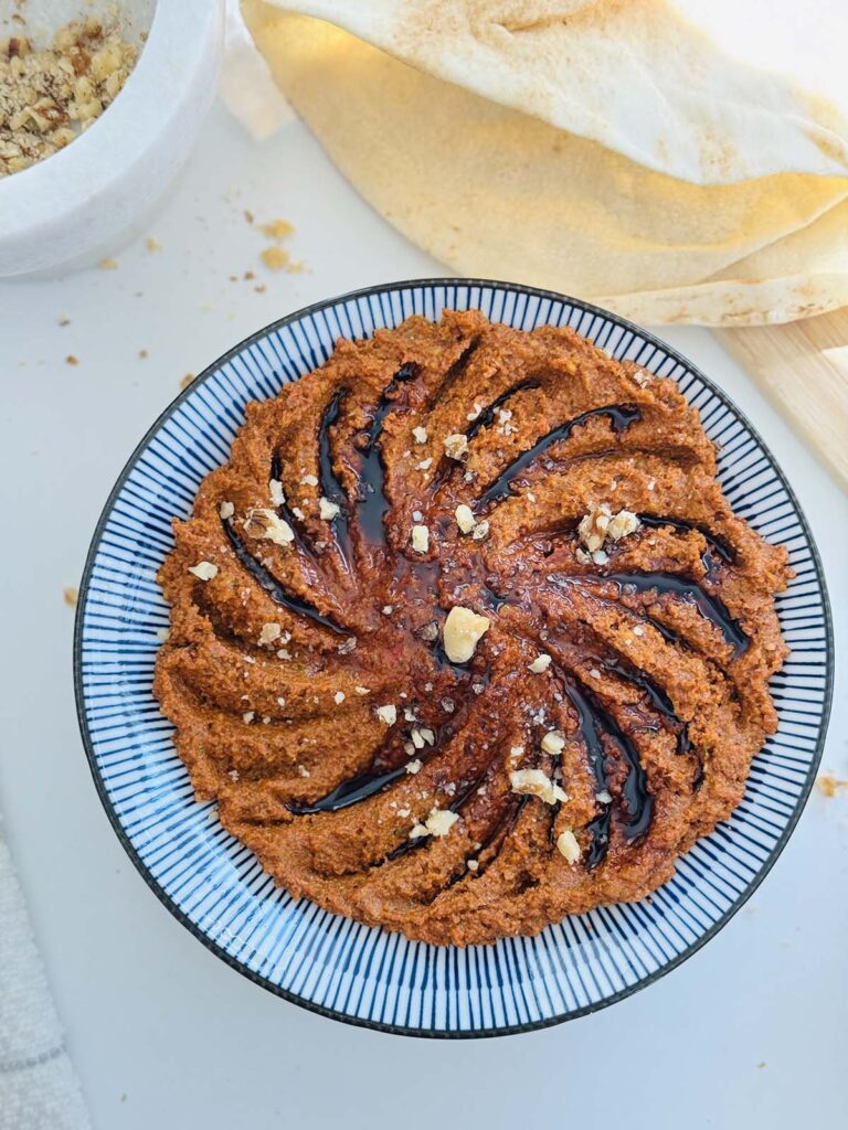 Muhamarra Roasted Red Pepper Dip in a round mezza platter with blue and white strips on top of a white countertop with a white marble mortar filled with crushed walnuts and pita bread on top of a wooden chopping board.