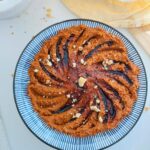 Muhamarra Dip in a round mezza platter with blue and white strips on top of a white countertop with a white marble mortar filled with crushed walnuts and pita bread on top of a wooden chopping board.