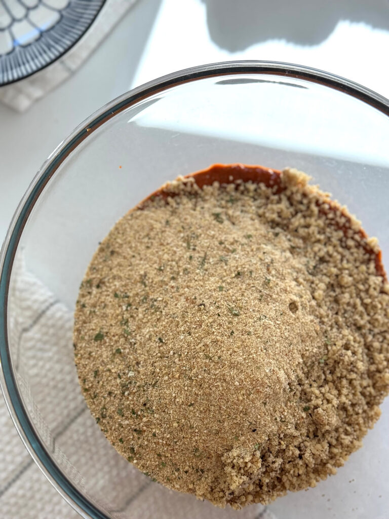 Glass mixing bowl filled with breadcrumbs, walnuts and peppers with a white stripped towel and blue and white pattern plate on a white tabletop. 