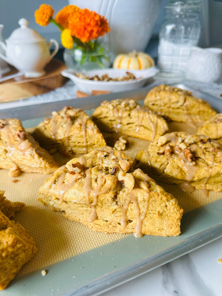 Maple Cinnamon Spice Tahini Scones Dessert in a silver metal baking pan with a rubber silicone mat with a grey accent fabric on table top with cinnamon sticks, and loose walnuts laying on top of grey lacey fabric with a white and orange spripped pumpkin and a white plate filled with walnuts and a glass bowl filled with cream