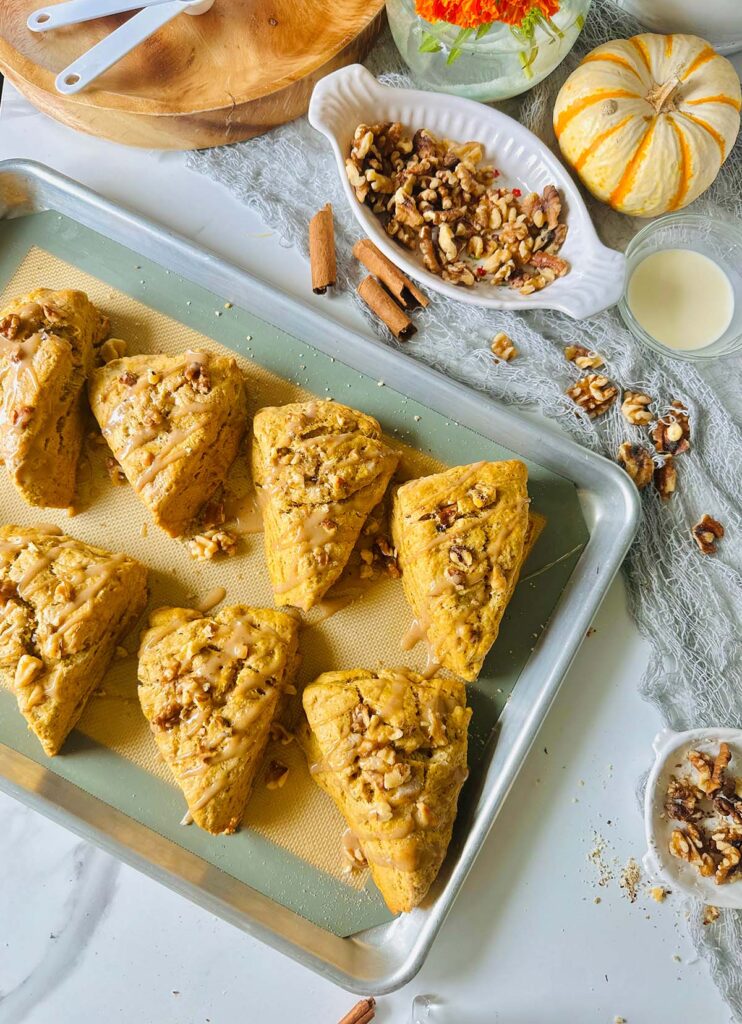 Maple Cinnamon Spice Tahini Scones Dessert in a silver metal baking pan with a rubber silicone mat with a grey accent fabric on table top with cinnamon sticks, and loose walnuts laying on top of grey lacey fabric with a white and orange spripped pumpkin and a white plate filled with walnuts and a glass bowl filled with cream