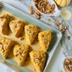 Maple Cinnamon Spice Tahini Scones Dessert in a silver metal baking pan with a rubber silicone mat with a grey accent fabric on table top with cinnamon sticks, and loose walnuts laying on top of grey lacey fabric with a white and orange spripped pumpkin and a white plate filled with walnuts and a glass bowl filled with cream