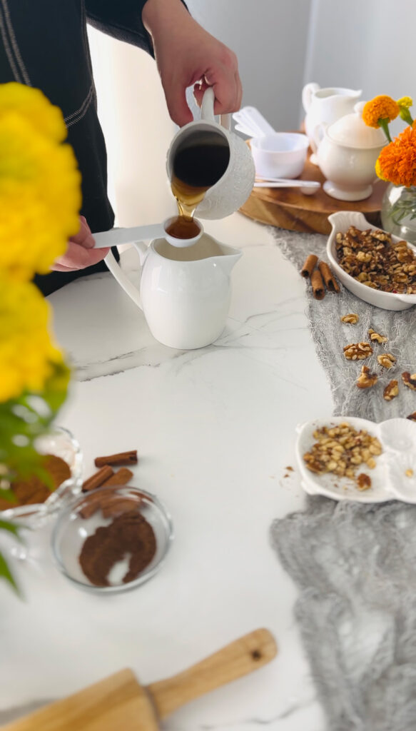 Maple Cinnamon Spice Tahini Scones Dessert white pitcher with a white measuring spoon pouring maple syrup into measuring spoon with a grey accent fabric on table top with cinnamon sticks, and loose walnuts laying on top of grey lacey fabric with a white and orange spripped pumpkin and a white plate filled with walnuts and a glass bowl filled with cream