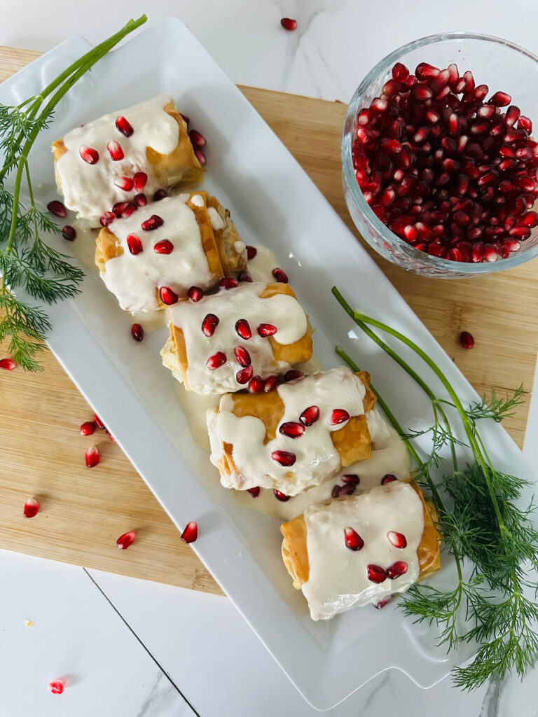 Crispy Phyllo w/ Eggplant Rolls with Tahini Sauce - Bite size phyllo dough bites with tahini sauce and pomegranates as a garnish on a white serving platter accented with fresh dil herbs and pomegranates in a glass bowl ontop of a wooden chopping board.