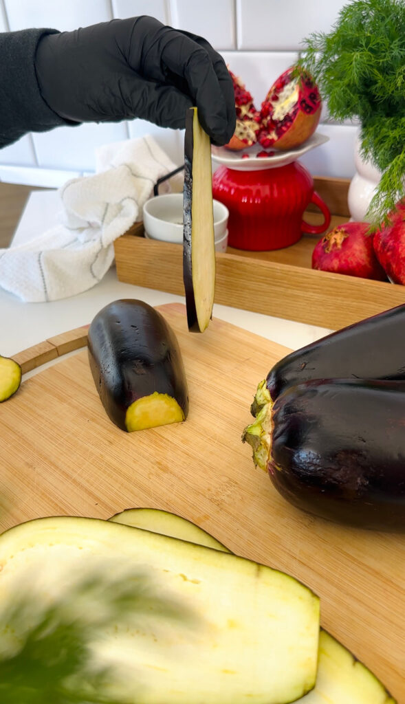 Crispy Phyllo Eggplant Rolls with Tahini Sauce - Eggplant slicing on a wooden chopping board with a wooden tray with a red stand with pomegranates and a white towel in the background eggplants on a wooden chopping board.