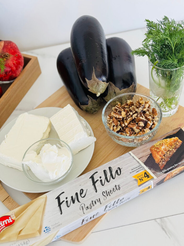 Crispy Phyllo Eggplant Rolls with Tahini Sauce - ingredients phyllo dough box that reads fine fillo dough, three eggplants cheese on a white platter with a bowl of walnuts a glass filled with fresh dill and a wooden tray of pomegranates on top of a wooden chopping board.