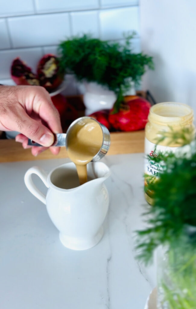 Crispy Phyllo Eggplant Rolls with Tahini Sauce - a white pitcher with a metal measuring cup pouring tahini sauce into a white pitcher with fresh dill and a jar of tahini in the background along with a wooden tray with pomegrates and a white vase filled with fresh dill.