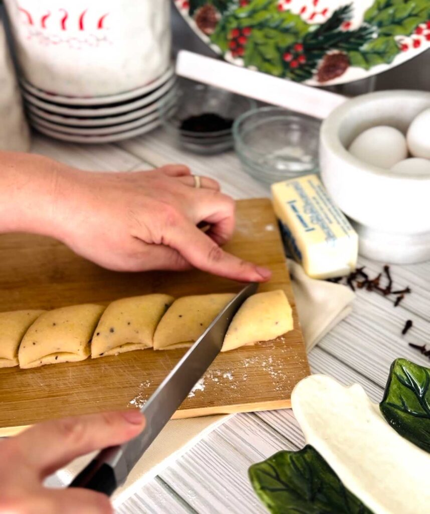 Ma'amoul Date Filled Cookies wooden board with rolled out dough formed into a log shape cutting with a knife into small pieces with a stick of butter, white bowl with eggs, small glass bowls with ingredients white canisters with whole cloves accenting the table with a ceramic platter on the bottom right corner with a town underneath wooden board.