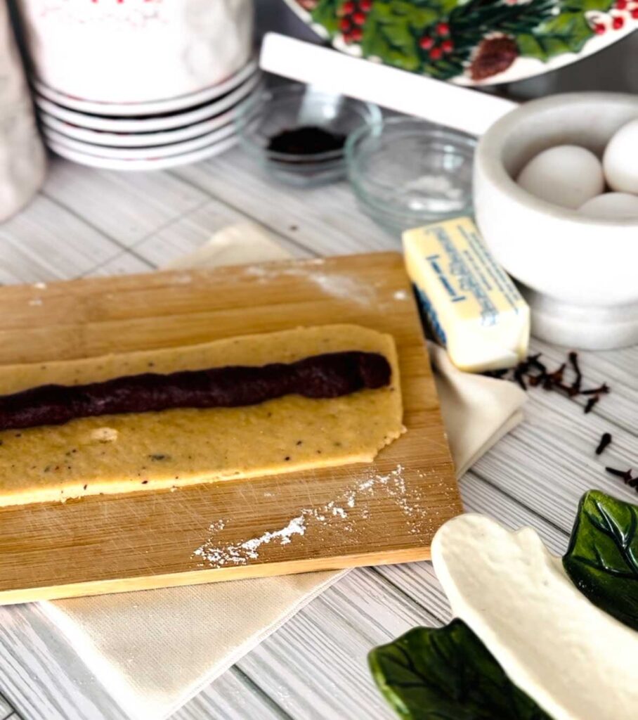 Ma'amoul Date Filled Cookies wooden board with rolled out dough with date paste in the center into a log shape with a stick of butter, white bowl with eggs, small glass bowls with ingredients white canisters with whole cloves accenting the table with a ceramic platter on the bottom right corner with a town underneath wooden board.