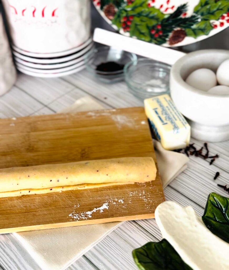 Ma'amoul Date Filled Cookies wooden board with rolled out dough formed into a log shape with a stick of butter, white bowl with eggs, small glass bowls with ingredients white canisters with whole cloves accenting the table with a ceramic platter on the bottom right corner with a town underneath wooden board.