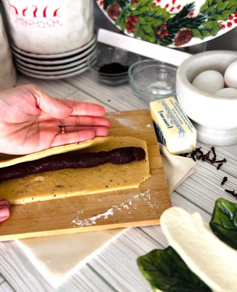 Ma'amoul Date Filled Cookies wooden board with rolled out dough with date paste in the center into a log shape and folding over dough with a stick of butter, white bowl with eggs, small glass bowls with ingredients white canisters with whole cloves accenting the table with a ceramic platter on the bottom right corner with a town underneath wooden board.