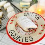 Ma'amoul Date Filled Cookies - Cookie on a christmas platter that reads santa's cookies with a cookie in the center with powdered sugar sprinkled over the top with a lit candle a stick of butter and cookies on a metal baking sheet in background.