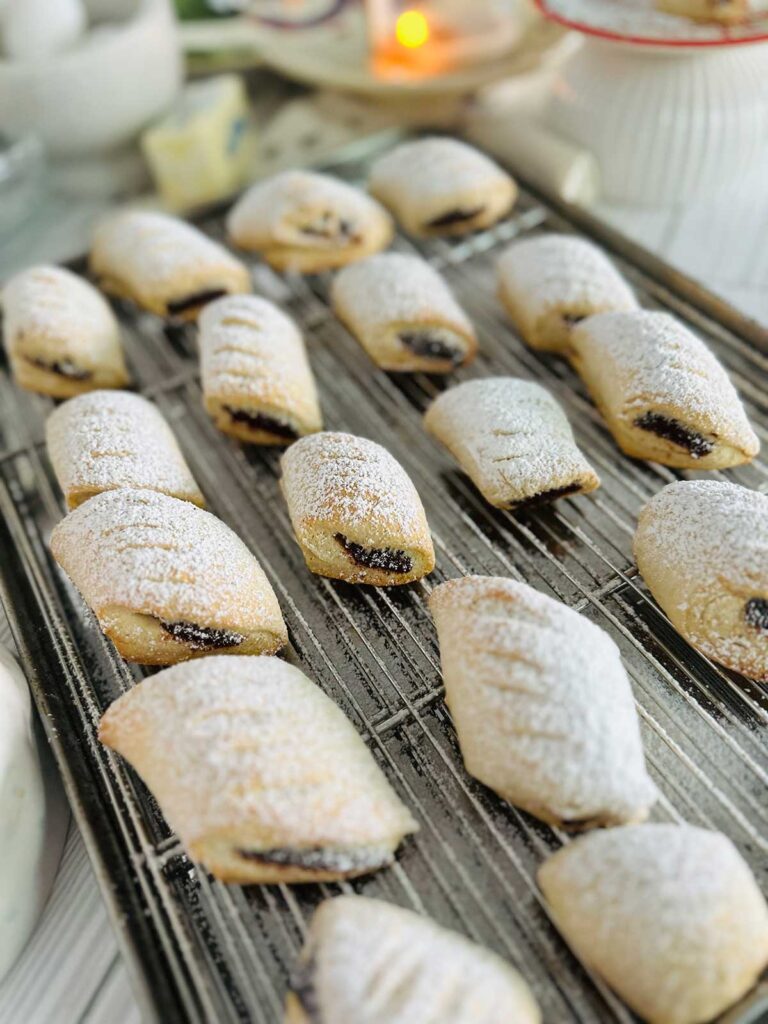 Ma'amoul Date Filled Cookies - Cookie on a christmas platter that reads santa's cookies with a cookie in the center with powdered sugar sprinkled over the top with a lit candle a stick of butter and cookies on a metal baking sheet in background.