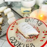 Ma'amoul Date Filled Cookies - Cookie on a christmas platter that reads santa's cookies with a cookie in the center with powdered sugar sprinkled over the top with a lit candle a stick of butter and cookies on a metal baking sheet in background.