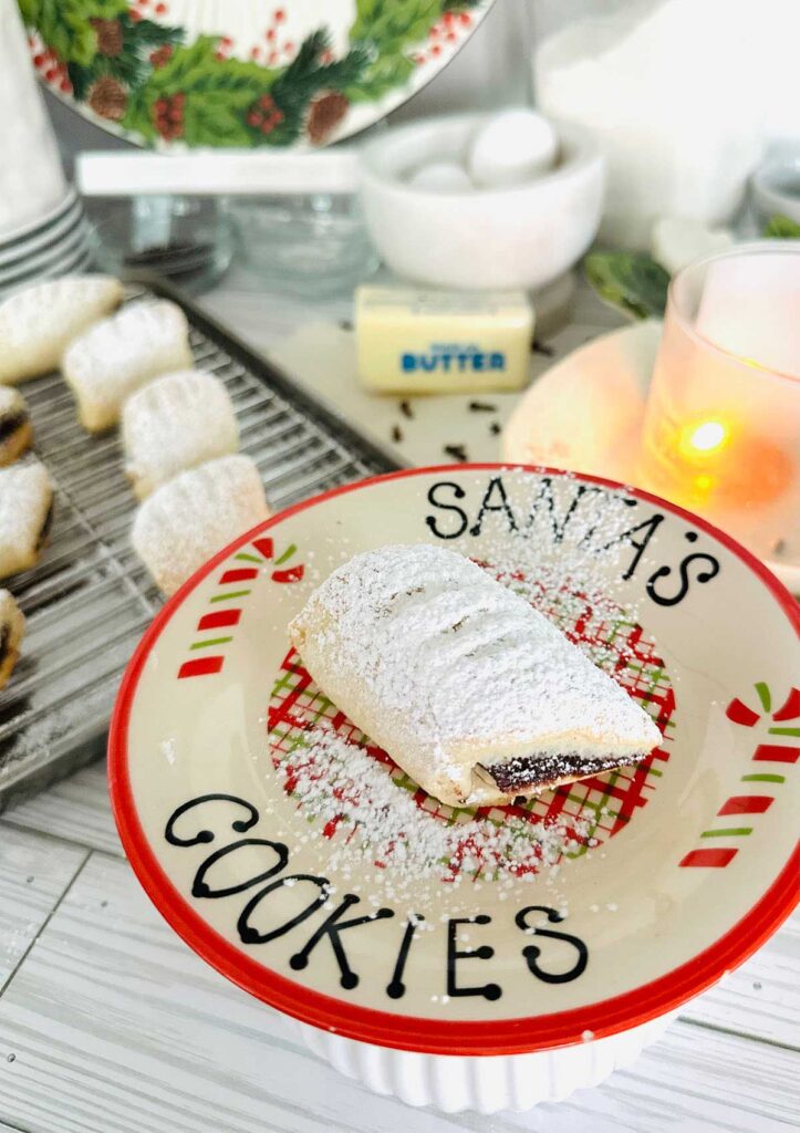 Ma'amoul Date Filled Cookies - Cookie on a christmas platter that reads santa's cookies with a cookie in the center with powdered sugar sprinkled over the top with a lit candle a stick of butter and cookies on a metal baking sheet in background.