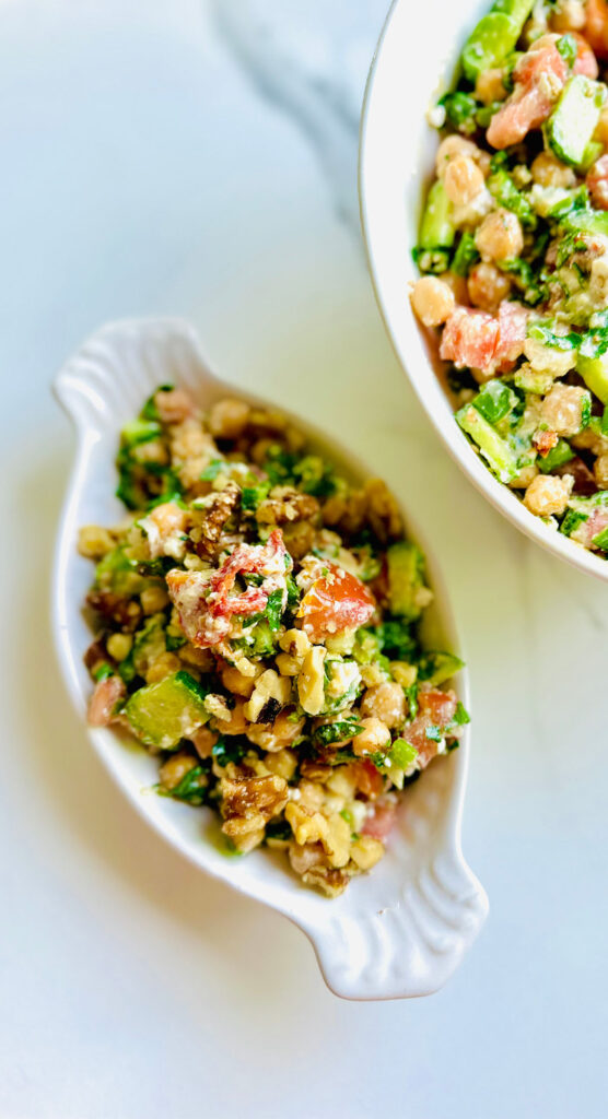 Chickpea Cucumber and Tomato Salad with Sun-dried Tomatoes in a white oval appetizer platter and a white mixing bowl filled with salad with a lit candle, potted plant in a grey planter and a black and white decorative sign saying Good Food in background ontop of a white countertop.