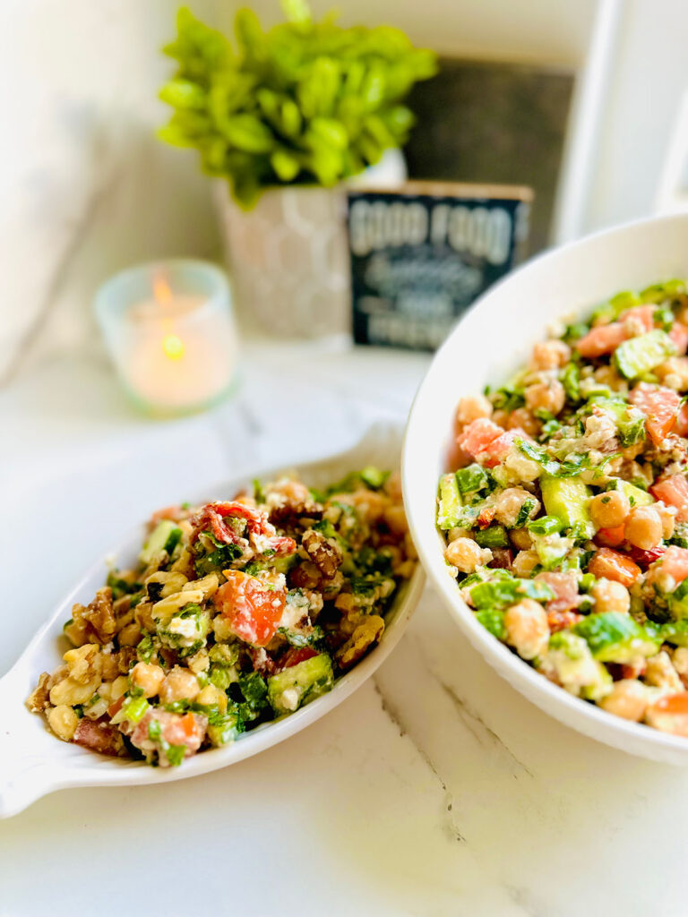 Chickpea Cucumber and Tomato Salad with Sun-dried Tomatoes in a white oval appetizer platter and a white mixing bowl filled with salad with a lit candle, potted plant in a grey planter and a black and white decorative sign saying Good Food in background ontop of a white countertop.