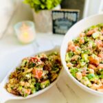 Chickpea Cucumber and Tomato Salad with Sun-dried Tomatoes in a white oval appetizer platter and a white mixing bowl filled with salad with a lit candle, potted plant in a grey planter and a black and white decorative sign saying Good Food in background ontop of a white countertop.