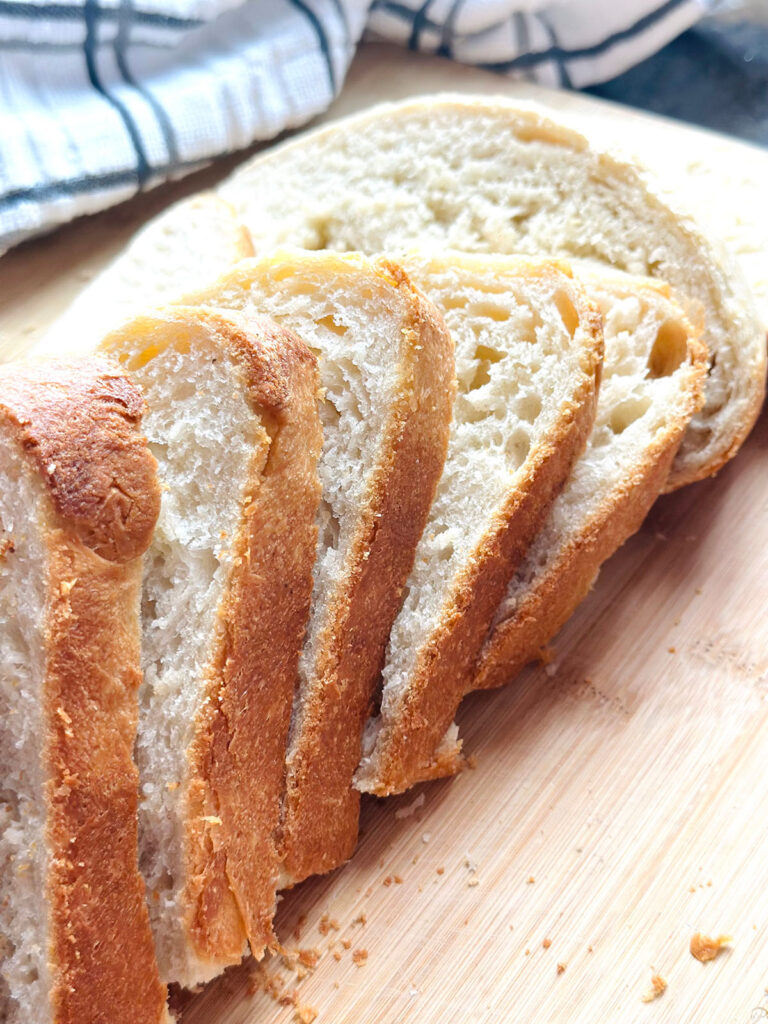 Homemade Fresh Bread Made Easy - Sliced loaf of bread ontop of a wooden chopping board with a white stipped kitchen towl and a bread knife on a black countertop.