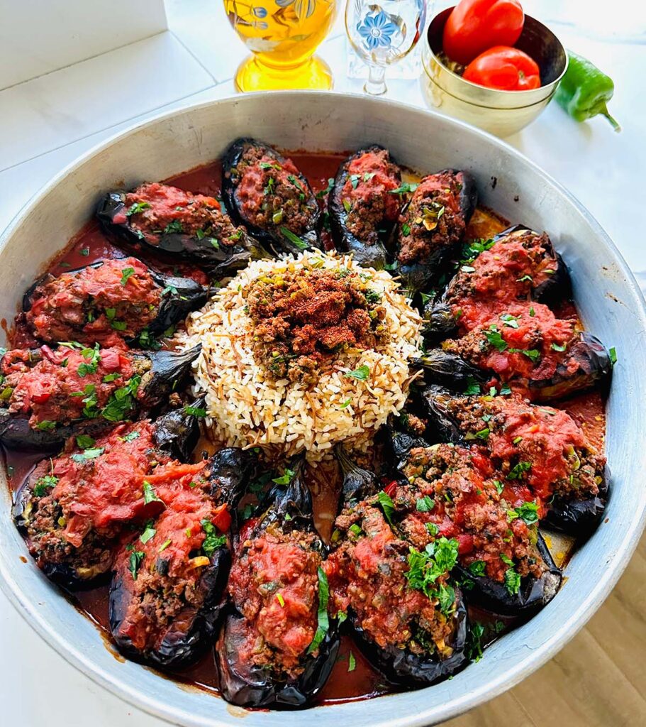 Karniyarik - Stuffed Eggplants - topped with tomato sauce and fresh herb with rice in the center in a round baking tin with a golden bowl of tomatoes and a glass oil pitcher with a cup on a white countertop