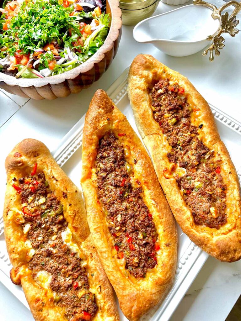 Middle Eastern Meat Flatbreads - Three flatbreads filled with meat on a white serving platter and a wooden bowl with a chopped salad with a white pitcher on a white countertop.