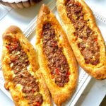 Middle Eastern Meat Flatbreads - Three flatbreads filled with meat on a white serving platter and a wooden bowl with a chopped salad with a white pitcher on a white countertop.