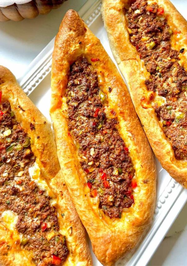 Middle Eastern Meat Flatbreads - Three flatbreads filled with meat on a white serving platter and a wooden bowl with a chopped salad with a white pitcher on a white countertop.