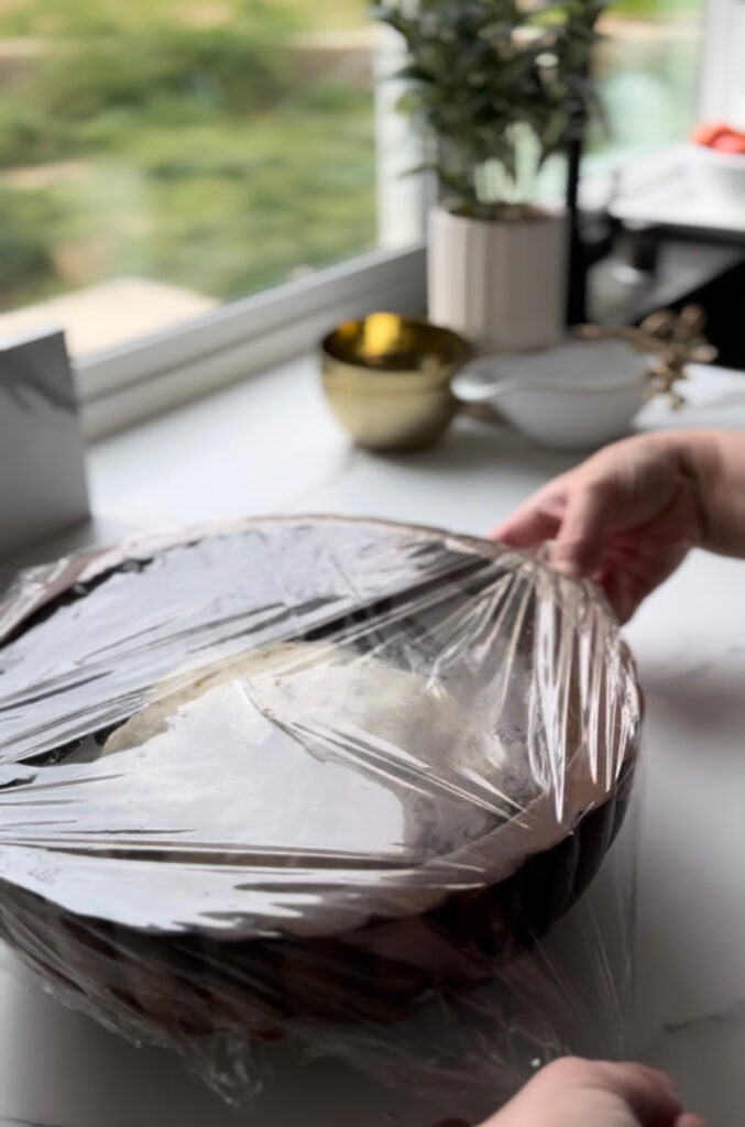 Middle Eastern Meat Flatbreads - wooden bowl with dough inside bowl with plastic wrap ontop with a wooden tray with white and glass bowls in background with a wooden chopping board on a white countertop.