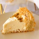 The Best No-Bake Kadayif/Kanafeh Cheesecake - a slice of cheesecake that has crushed pistachios on a white plate, with syrup being over the cheesecake from a white container. A whole cheesecake on a white stand with golden feet with a cross of the center of the cheesecake in the background.