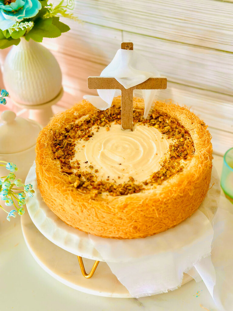 The Best No-Bake Kadayif/Kanafeh Cheesecake - Kadayif Cheesecake placed on a white stand with gold feet. Cross with white cloth placed on top of cheesecake. White vase with blue flowers in the background.