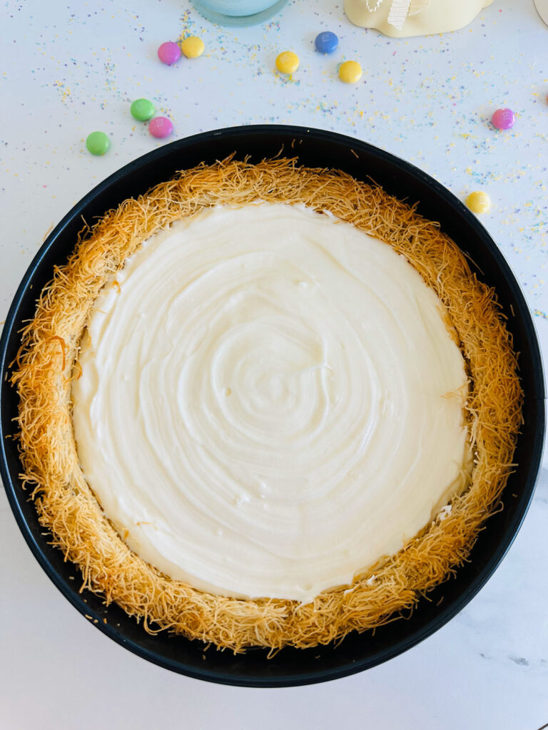 The Best No-Bake Kadayif/Kanafeh Cheesecake - Top view of cheesecake in springform pan. White cream swirl  in the center with golden cripsy Kadayif base. Colored M&M's scattered on countertop arond pan.