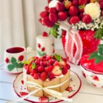 Strawberry / Raspberry Trifle Cake with Puff Pastry - Strawberry themed photo with a red strawberry vase filled with fresh strawberries with white roses with a white laced bow fresh mint, strawberry cup, strawberry sugar container, with fresh raspberries, strawberries on top of cake that has graham crackers around with a white with a golden ribbon around cake.