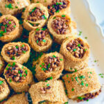 Jalapeño Phyllo Poppers with Roasted Pine Nuts - Cheese stuffed with crispy phyllo dough rolled in sesame seeds and baked till crispy and topped with roasted pine nuts and fresh chives ontop of a white serving platter.