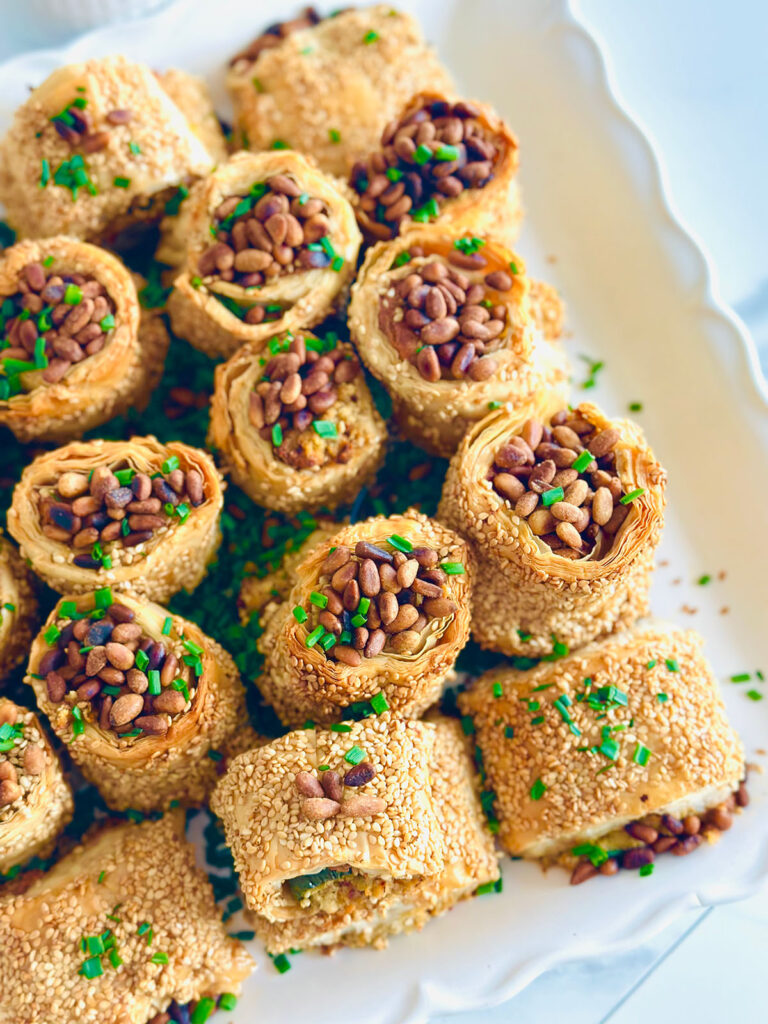 Jalapeño Phyllo Poppers with Roasted Pine Nuts - Cheese stuffed with crispy phyllo dough rolled in sesame seeds and baked till crispy and topped with roasted pine nuts and fresh chives ontop of a white serving platter.
