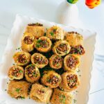 Jalapeño Phyllo Poppers with Roasted Pine Nuts - Cheese stuffed with crispy phyllo dough rolled in sesame seeds and baked till crispy and topped with roasted pine nuts and fresh chives ontop of a white serving platter. White vase with one single tulip as decore on a white table top.
