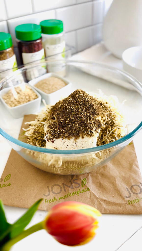 Jalapeño Phyllo Poppers with Roasted Pine Nuts - Cheese stuffed with crispy phyllo dough rolled in sesame seeds and baked till crispy in white muffin tin with Jons Marketplace seasonings and paper under glass bowl filled with cheese filling and seasonings with two white square containers filled with sesame seeds and pine nuts in background with a white pitcher filled with fresh tulips.