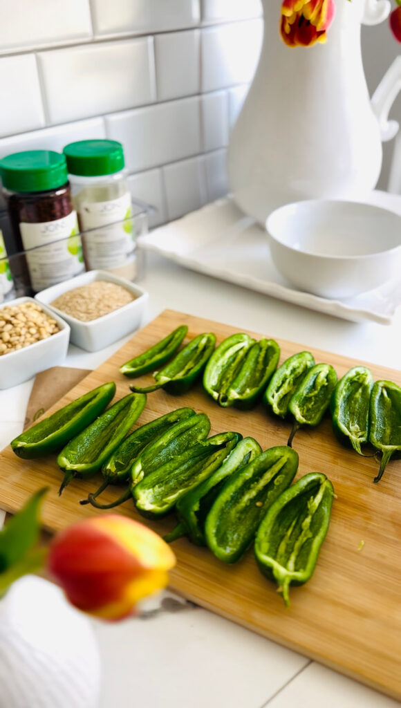 Jalapeño Phyllo Poppers with Roasted Pine Nuts - cut jalapenos on a wooden board with a white pitcher in background with tulips, seasonings with green lids in background with white square containers with sesame seeds and pine nuts in them with a white empty bowl on the top right.
