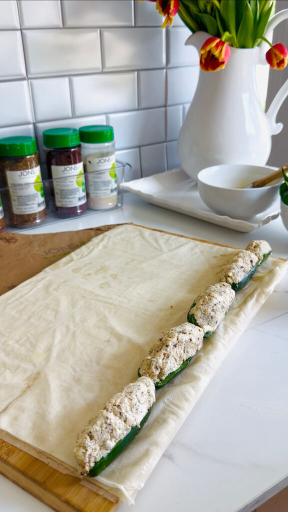 Jalapeño Phyllo Poppers with Roasted Pine Nuts - wooden chopping board with phyllo dough laying on flat surface with stuffed jalapenos on the edge of the dough. Seasonings in backgound with green lids, white pitcher with fresh tulips ontop of a white platter for decor.