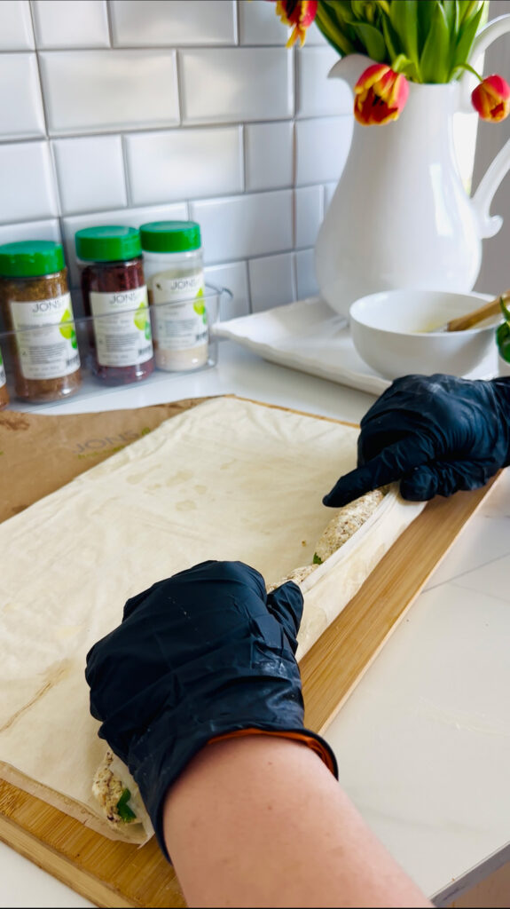 Jalapeño Phyllo Poppers with Roasted Pine Nuts - Rolling phyllo dough over with two hands with black gloves. Seasonings with green lids in background. White pitcher vase filled with fresh tulips. So Yummy and Delicious! #appetizer #phyllodoughbites #middleeastern #jalapenopoppers| galoreofflavors.com