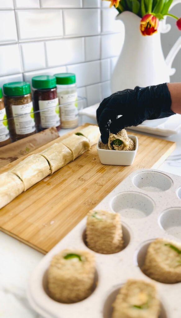 Jalapeño Phyllo Poppers with Roasted Pine Nuts - wooden chopping board with phyllo dough cut rolls with a white muffin tin filled with cut poppers. Black glove rolling popper into sesame seeds in a white container, seasonings in backgound with green lids, white pitcher with fresh tulips ontop of a white platter for decor.