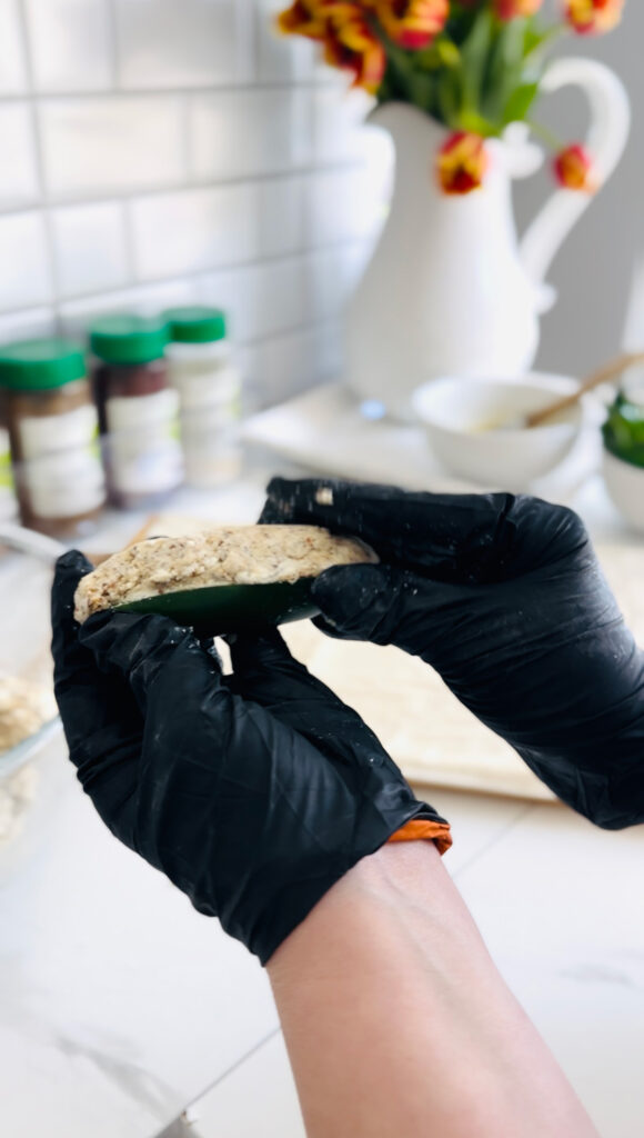Jalapeño Phyllo Poppers with Roasted Pine Nuts - jalapeno half stuffed with cheese holdig with two hands with black gloves with seasonings in background with green lids and white pitcher with fresh tulis inside with white bowl in background.