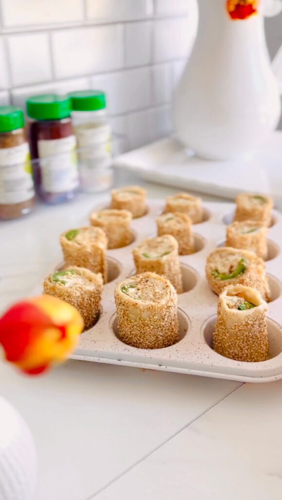 Jalapeño Phyllo Poppers with Roasted Pine Nuts - Cheese stuffed with crispy phyllo dough rolled in sesame seeds and baked till crispy in white muffin tin with Jons Marketplace seasonings in background with a white pitcher filled with fresh tulips.