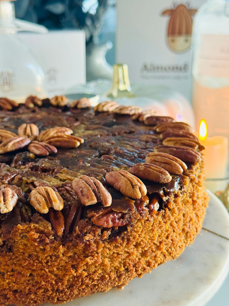 Buttery Pecan Upside Down Cake - Almond Cow - Pecan upside cake cut slice on white plate with fall docor and a lit candle in background with a white pumpkin decor in background.
