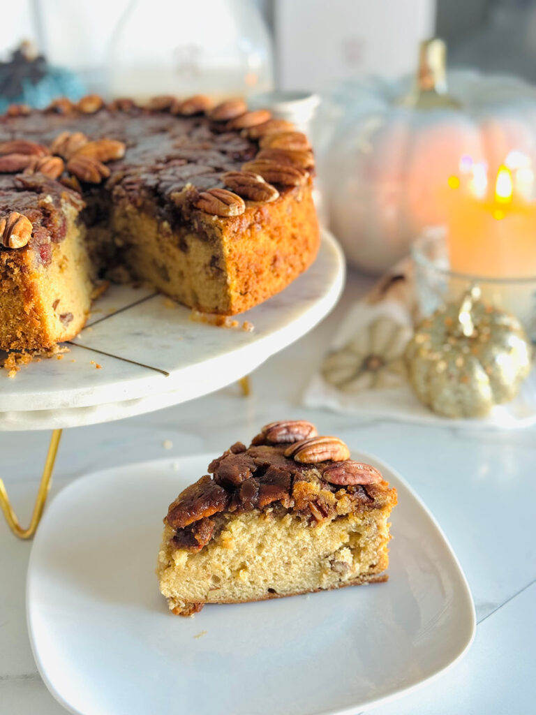 Buttery Pecan Upside Down Cake - Almond Cow - Pecan upside cake cut slice on white plate with fall docor and a lit candle in background with a white pumpkin decor in background.